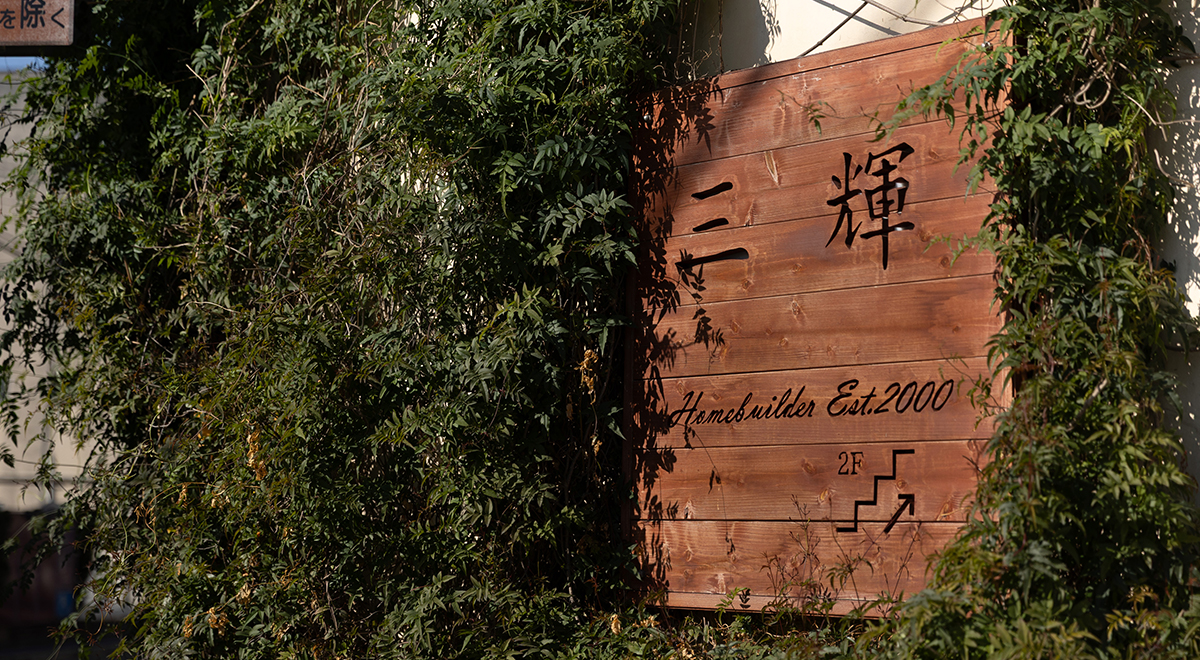 「三輝」の看板が掲げられた建物の前で、作業着姿のスタッフを含む7人の社員が並んで写る集合写真